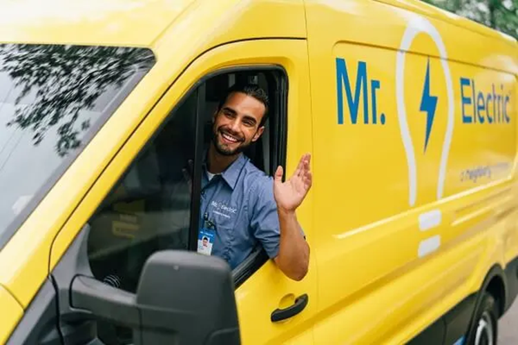 A Smiling Mr. Electric Electrician in a Yellow Mr. Electric Van Waves His Arm Out of the Open Driver-Side Window
