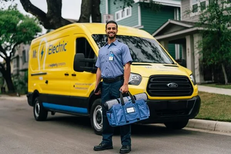 A Smiling Mr. Electric Electrician Stands in Front of a Mr. Electric Van Holding a Bag with a Rolled Door Mat on Top of It 