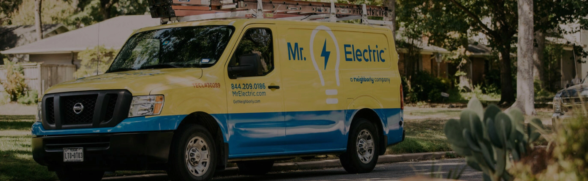 A Smiling Mr. Electric Electrician Stands in Front of a Mr. Electric Van Holding a Bag with a Rolled Door Mat on Top of It 