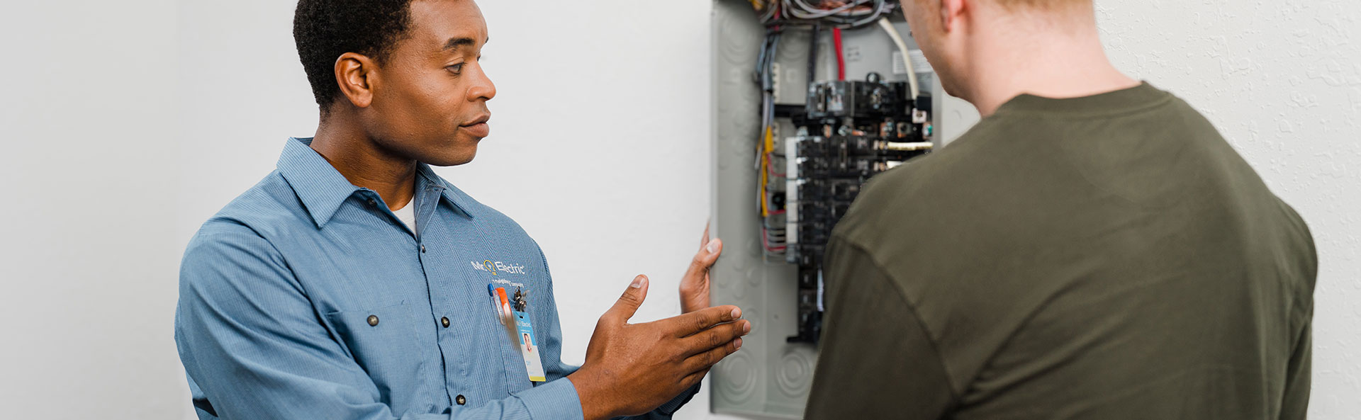 A Mr. Electric Service Professional Holding a Tablet Points at an Open Electrical Panel While Talking to a Smiling Man 