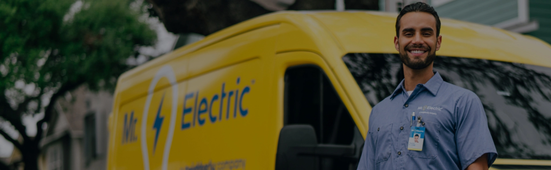 A Smiling Mr. Electric Electrician Stands in Front of a Mr. Electric Van Holding a Bag with a Rolled Door Mat on Top of It 