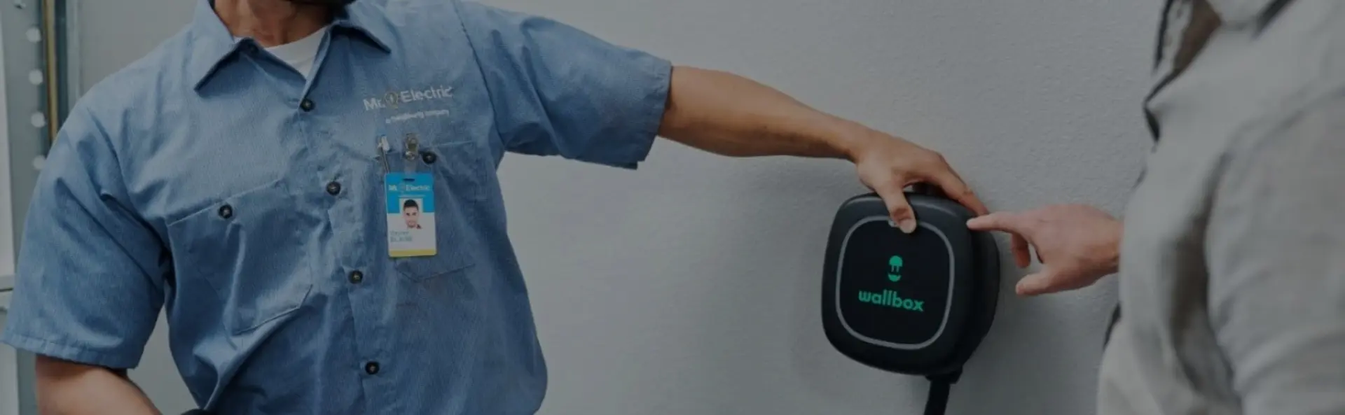 A smiling Mr. Electric electrician touches an EV charger on the wall to show another smiling man watching how to use it .